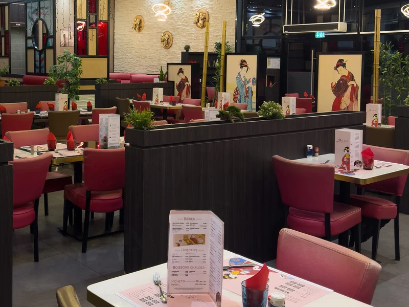 Salle vide avant le service : tables dressées avec des serviettes rouges, chaises bordeaux, claustras en bois sombre, tableaux japonais au fond et suspensions lumineuses